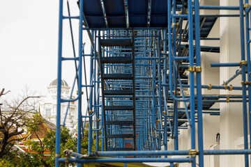 Complex network of blue scaffolding is set up against building construction, with white domed structure visible background. scene captures intricate geometry and industrial feel of construction