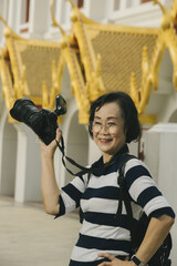 Smiling woman holding camera stands in front of building with ornate golden architecture, wearing striped shirt and glasses. scene conveys sense of joy and exploration