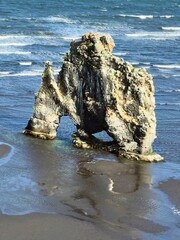 A dramatic basalt rock formation, resembling a sea stack, in the middle of a calm sea with a small beach and distant mountains, ideal for themes of natural wonder and secluded coastal landscapes.