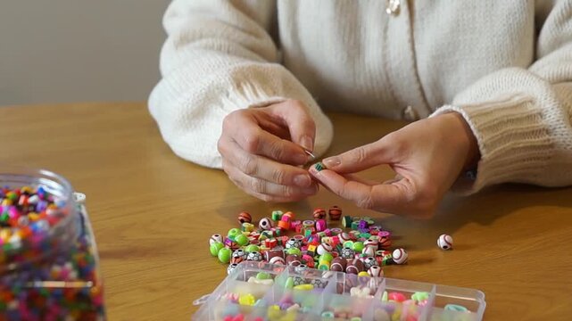 A detailed shot of hands carefully placing a fruit-shaped bead onto a needle and thread, part of a creative DIY jewelry workshop at home.