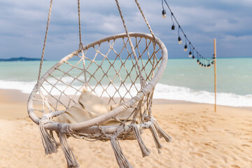 Empty hanging chair on sandy tropical beach with ocean waves and string lights, calm seaside atmosphere, summer travel concept