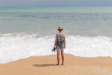 Young woman standing barefoot on sandy beach facing ocean waves, wearing hat and backpack, travel and freedom concept with wide sea horizon 