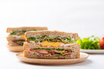 Whole wheat bread sandwich with bacon, lettuce, tomato and fried egg on wooden plate with white background