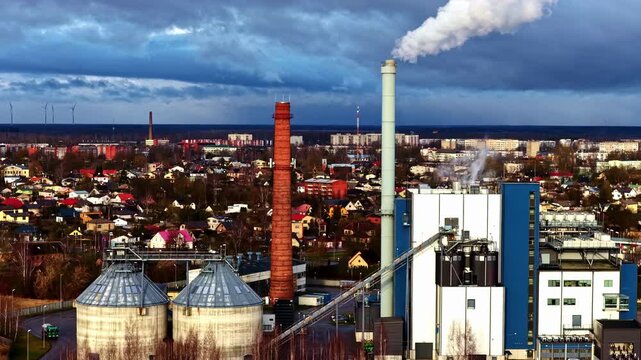Drone shot circling industrial plant with steam emitting from red brick stacks against winter city skyline in Jelgava