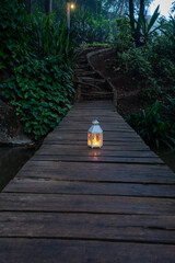 A glowing white vintage lantern placed on a wooden boardwalk path in a lush tropical garden at...