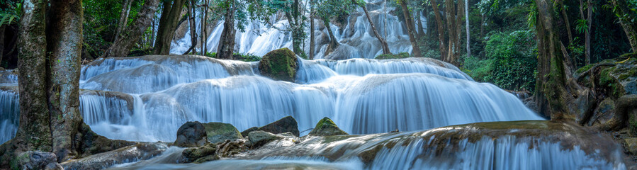 Fototapeta premium Beautiful horizontal landscape of a tiered limestone waterfall flowing through a lush green tropical forest. Serene nature scenery with silk water effect and trees framing the cascading stream.