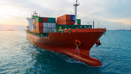Close-up front view of a large red cargo ship sailing in the open sea at sunset. Loaded with colorful shipping containers, representing global logistics and international trade transportation.