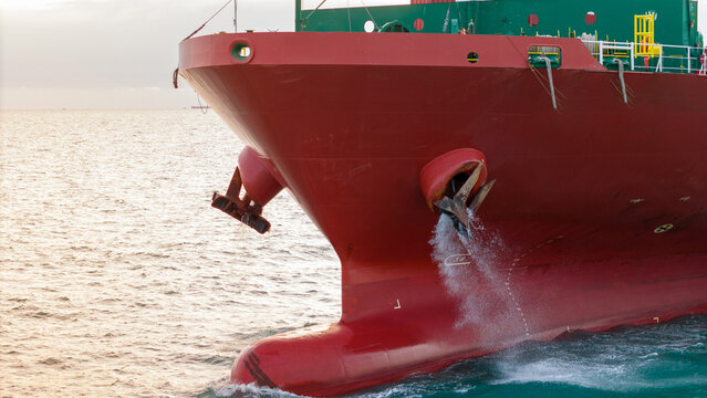 a red cargo ship's bow sailing in the sea. Water splashing from the anchor housing area, highlighting maritime transport operations and heavy industrial machinery mechanics.