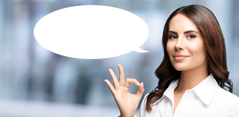 Portrait image - happy smiling business woman wear white shirt show point ok okay hand sign gesture. Businesswoman standing on blur office background with blank free space mockup speech bubble