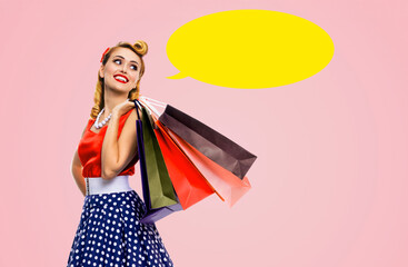 Black Friday Sales concept - beautiful cute blond woman in pinup pin-up dress holding carrying colourful shopping bags isolated pink background with yellow empty mockup speech bubble. Mock up ad image