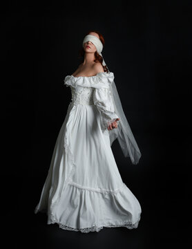 full length portrait beautiful female model wearing antique white historical ghost bride wedding dress gown, veiled mask blindfold covering face. isolated walking figure on dark studio background.