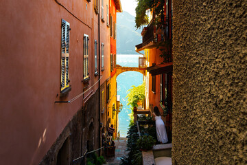 Naklejka premium Locals enjoy the water views from a narrow colorful alley with an archway leading to the shores of Lake Como in the lakefront resort village of Varenna.