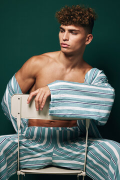 Young man with curly hair wearing striped pajama pants and matching shirt with open chest sits sideways on a white chair against dark green background with pensive expression.