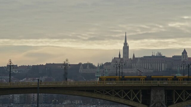 Yellow Tram Crossing Margaret Bridge With Ornate Streetlamps, Matthias Church Tower In Background Against Overcast Sky. wide shot