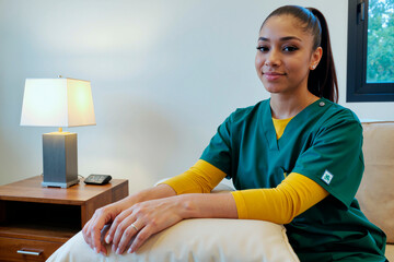 Portrait of young adult Hispanic woman sitting on sofa wearing medical scrubs, looking into camera with slight smile, hands resting on pillow, modern interior background visible