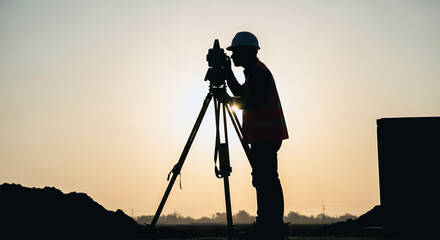 Construction site surveyor engineer silhouette operating total station instrument against a bright sunset sky.
