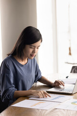 Young Asian project manager woman analyzing paper sales reports at laptop, doing marketing research...