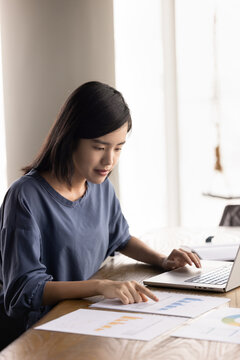 Young Asian project manager woman analyzing paper sales reports at laptop, doing marketing research for business project development, working on strategy of Internet product promotion. Vertical shot