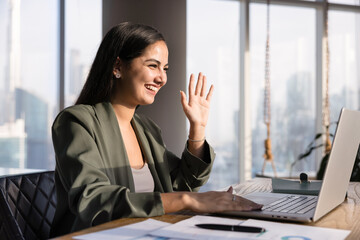 Cheerful young Hispanic business professional woman waving hand hello at laptop screen, using computer for online remote work communication, speakin on video conference chat