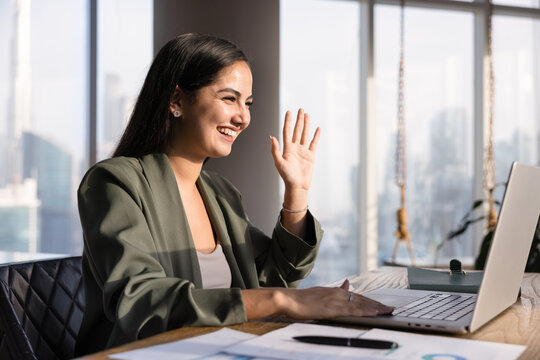 Cheerful young Hispanic business professional woman waving hand hello at laptop screen, using computer for online remote work communication, speakin on video conference chat