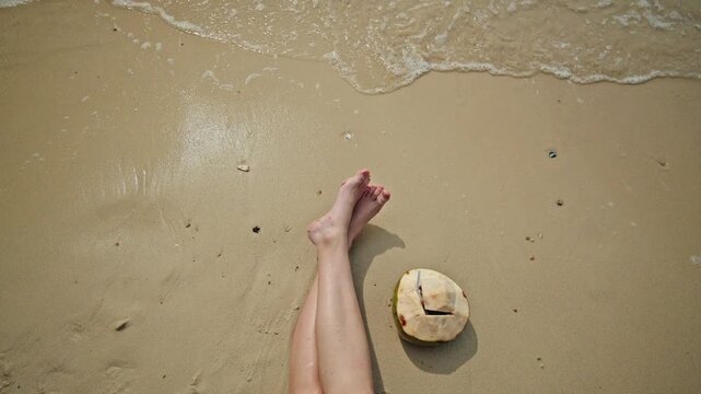 Young woman relaxing on sandy beach with bare legs and fresh coconut near gentle sea waves, tropical vacation mood with copy space