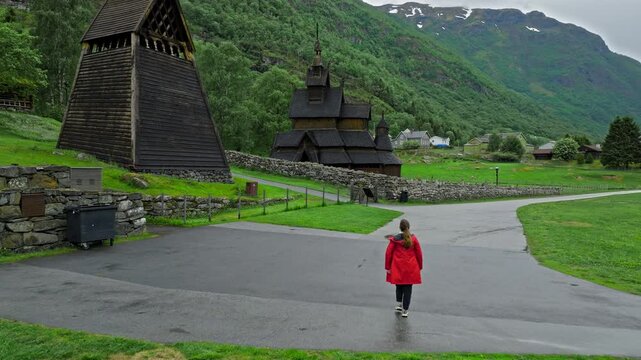 ourist walking toward Borgund Stave Church in a green mountain valley. Historic wooden church and bell tower stand behind stone walls under cloudy Norwegian skies.