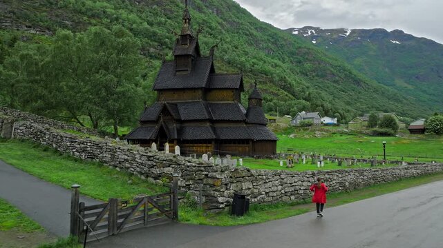 Tourist walking toward Borgund Stave Church on a quiet mountain day. Historic wooden church, bell tower, and stone walls sit in a green Norwegian valley under cloudy skies.