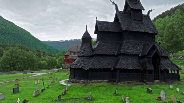 View of Borgund Stave Church surrounded by a historic cemetery. Dark wooden church stands in a green mountain valley under cloudy skies.