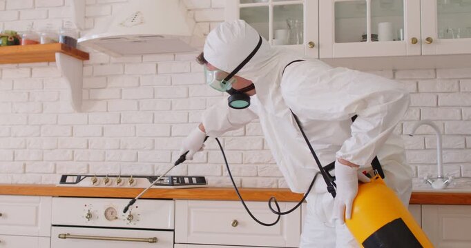 Exterminator in mask and goggles sprays insecticide indoors. Worker in respirator uses sprayer to disinfect kitchen surfaces, removing insects. Concept: home hygiene, safety, pest control services
