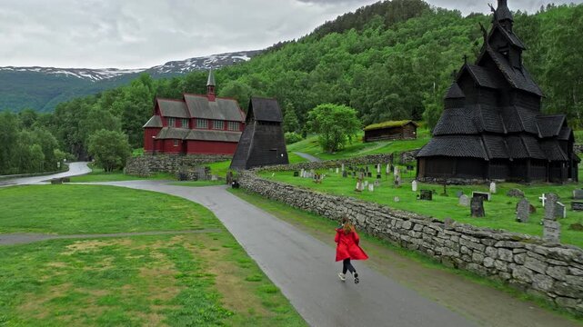 ourist running toward Borgund Stave Church in Norway surrounded by green mountains. Woman visits historic wooden stave church and cemetery create a peaceful cultural travel scene on a cloudy day.