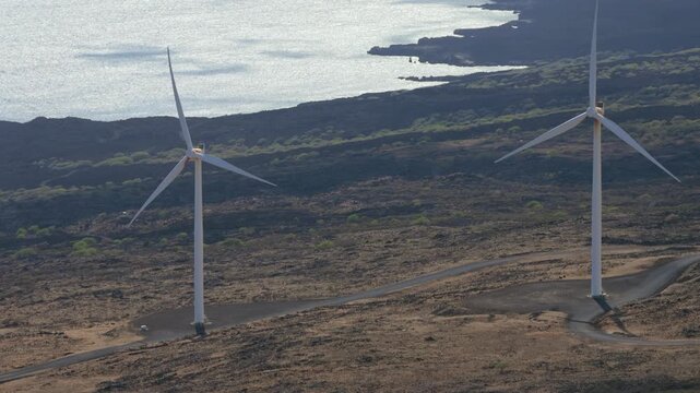 A high view captures white wind turbines on a rugged rocky coast. Sparkling ocean water and dark volcanic earth define this clean energy scene in a remote landscape. Shot on the Maui Coast.