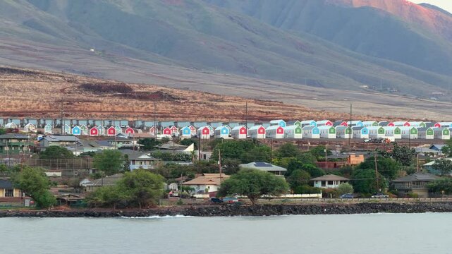 A drone captures a row of colorful modular housing units in Lahaina, Maui. The West Maui Mountains rise in the background, showing the landscape after the destructive 2023 wildfires.