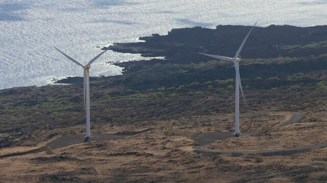 A high angle view shows white wind turbines on a dark rocky coast. The blue ocean and rugged volcanic landscape create a clean energy scene in a remote and natural environment. Shot on the Maui Coast