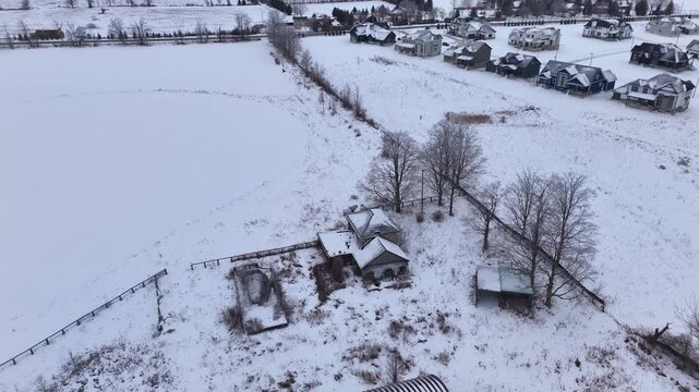 High-angle drone footage reveals a secluded farmhouse and outbuildings nestled in a vast, snow-covered field, bordering a residential development on a quiet winter day.