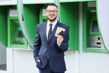 Young businessman with euro banknotes and credit card against ATM outdoors