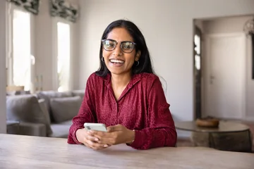 Fotobehang Ridders Cheerful beautiful young Indian smartphone user woman in trendy glasses holding mobile phone, looking at camera with toothy smile, laughing, posing for head shot portrait at home table  © fizkes