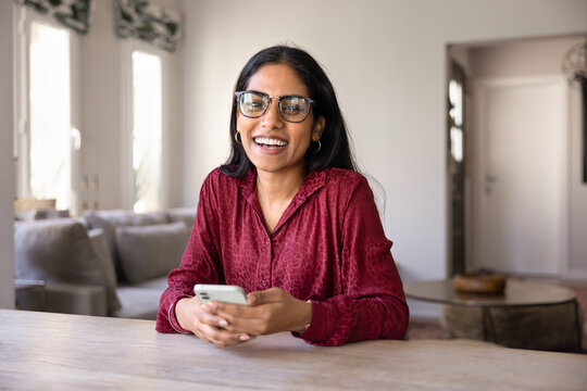 Cheerful beautiful young Indian smartphone user woman in trendy glasses holding mobile phone, looking at camera with toothy smile, laughing, posing for head shot portrait at home table
