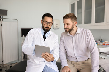 Health report review. Confident male doctor physician general practitioner in white coat hold digital tablet explain young man patient medical data lab test results on consultation in examination room © fizkes