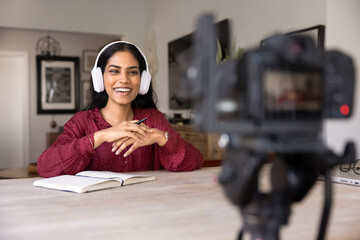 Happy young Indian influencer woman in wireless headphones using reflex camera on tripod for...