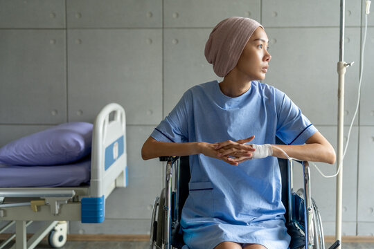 A cancer patient sits in a wheelchair with an IV drip looking stressed and sad. Asian Woman receiving IV therapy in a wheelchair showing visible signs of stress and depression