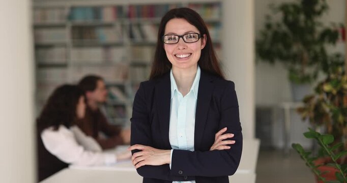 Professional female office employee or teacher posing, looking at camera with arms crossed standing in library, colleagues in background. Company staff member portrait, career growth and confidence