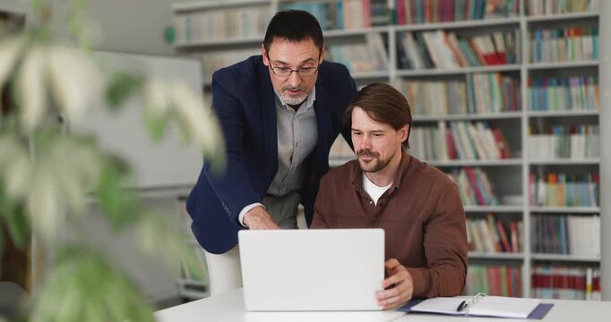 Young adult and middle-aged business men reviewing project data on laptop. Mentor helping with task completion, explain new corporate software to male colleague apprentice during teamwork in office