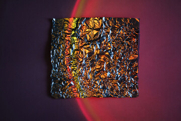 Crumpled metallic foil square on abstract neon gradient background with red light arc