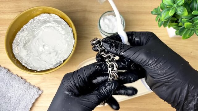 Close-up of black-gloved hands cleaning tarnished silver jewelry with a brush and baking soda on a wooden table. Concept of care and maintenance.