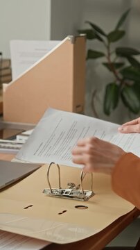 Vertical close view of office employee inserting printed documents into folder while organizing paperwork at workplace desk in contemporary office setting