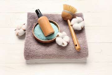 Bottle of cosmetic product, sea salt, massage brush, clean towel and cotton flowers on light wooden background