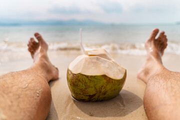 Man relaxing on sandy beach with fresh green coconut drink between legs, calm sea and distant mountains creating peaceful tropical vacation scene