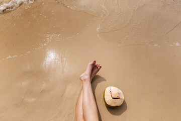 Young woman relaxing on tropical beach with fresh coconut drink on wet sand, gentle waves and warm sunlight creating calm summer vacation atmosphere with copy space