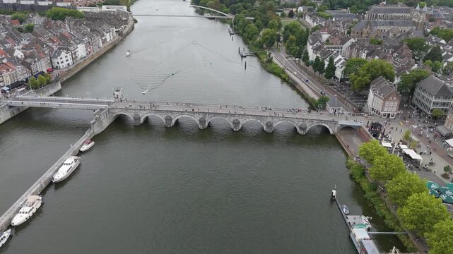 Drone footage of the historic Sint Servaasbrug, a medieval stone arch bridge spanning the Maas River in Maastricht. Surrounding views include the old town, waterfront, boats, and classic European arch
