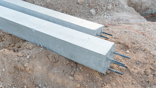 Pair of long square precast concrete pillars resting horizontally on a mound of excavated earth at a construction site. Pillars have exposed steel rebar ends at the front.